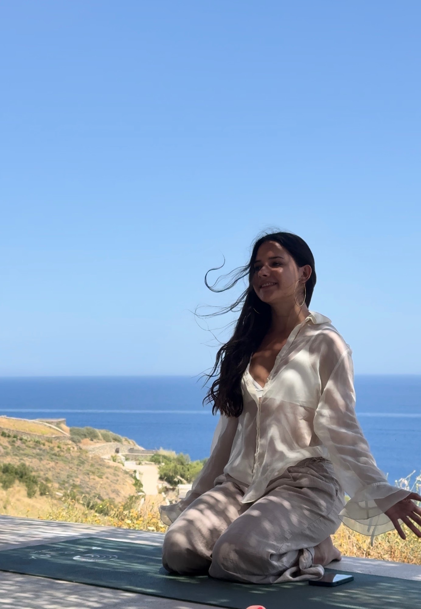 Mariana kneeling on a mat on a terrace above the sea, smiling, hair in the wind